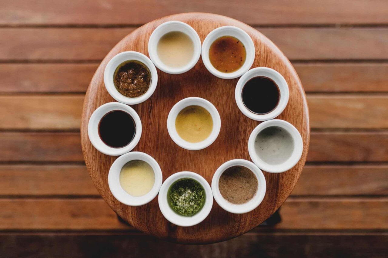 An overhead view of a round wooden serving board arranged with ten small white ramekins, each filled with a different sauce or condiment ranging from dark soy to green pesto and yellow mustard.