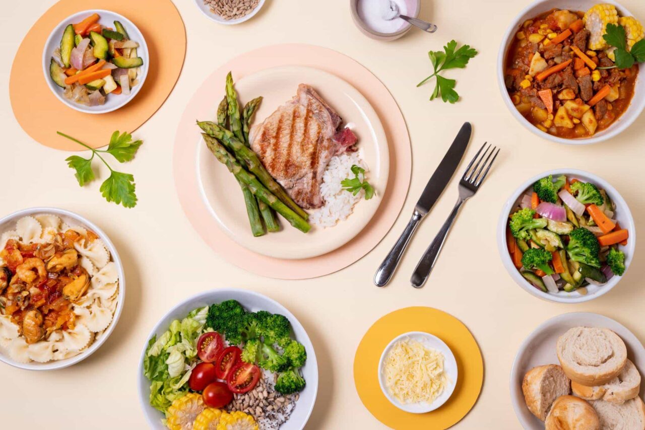 An overhead photograph of a variety of prepared meals on a light background, including a grilled steak with asparagus, seafood pasta, beef stew, vegetable salads, and bread rolls.