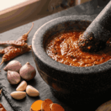 A close-up view of a heavy stone mortar and pestle containing a freshly ground red spice paste, surrounded by raw ingredients including dried chilies, shallots, garlic cloves, turmeric slices, lemongrass stalks, and whole spices like star anise and cinnamon on a dark slate surface.