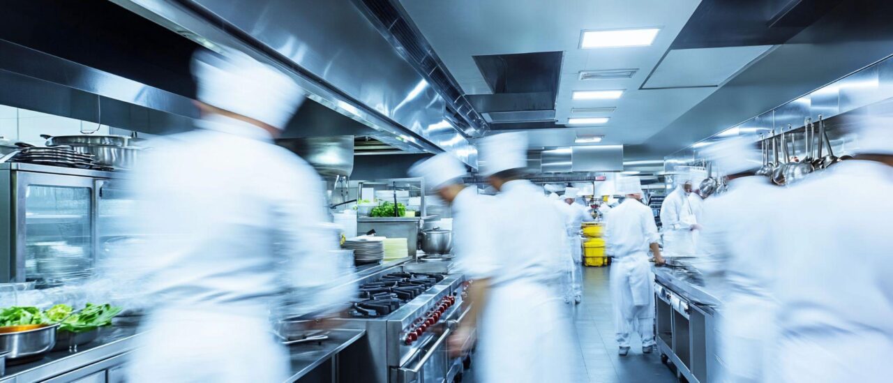 A wide shot of a bustling professional kitchen with several chefs in white uniforms moving quickly, creating a motion blur effect. The kitchen features stainless steel surfaces, industrial stoves, and hanging cookware.