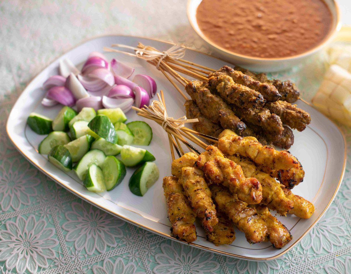 A white rectangular plate featuring several skewers of grilled chicken and beef satay. The meat is well-charred and served alongside fresh cucumber chunks, red onion wedges, and a side bowl of thick, peanut dipping sauce.