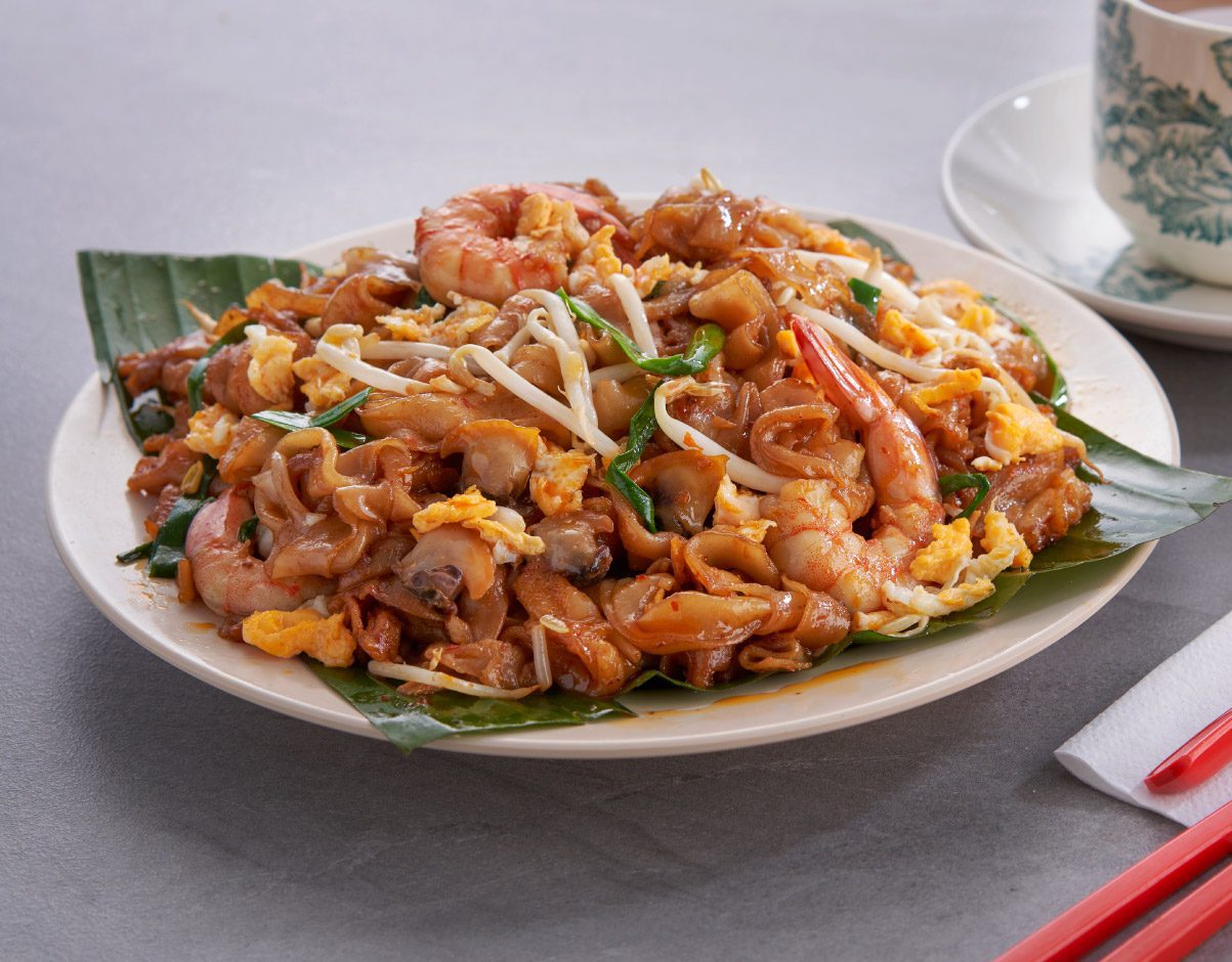 A close-up of a plate of Char Kway Teow, showing stir-fried flat rice noodles with large shrimp, scrambled eggs, bean sprouts, and green chives. The dish is served on a green banana leaf over a white plate.