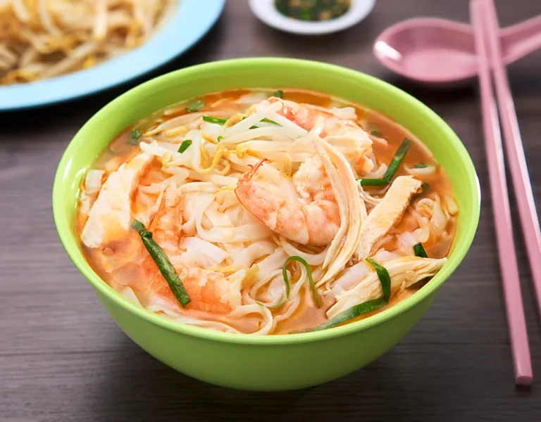 A green bowl filled with noodle soup containing flat rice noodles, sliced chicken, prawns, and bean sprouts in a light broth, accompanied by pink utensils.