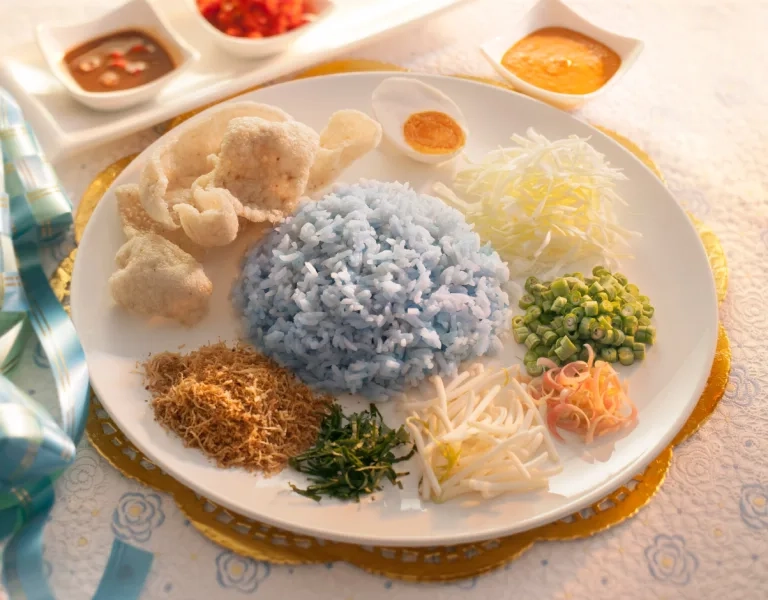 A traditional Malaysian Nasi Kerabu featuring a mound of blue-tinted rice in the center. It is surrounded by various accompaniments: fish crackers, salted egg, shredded cabbage, chopped long beans, herbs, and small bowls of spicy sambal and coconut-based sauce.