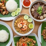 An overhead view of a wooden table spread with various Thai dishes served on white plates with green banana leaves. The meal includes shrimp fried rice, basil pork with rice, noodle dishes, and bowls of beef soup with meatballs.